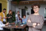 Eine Selbstbewusste Frau mit Brille steht lächelnd im Vordergrund eines modernen Büros, während im Hintergrund ein Team aus Kolleginnen und Kollegen gemeinsam arbeitet.